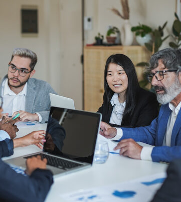 Group of people in business clothing sit around a table with laptops and paperwork