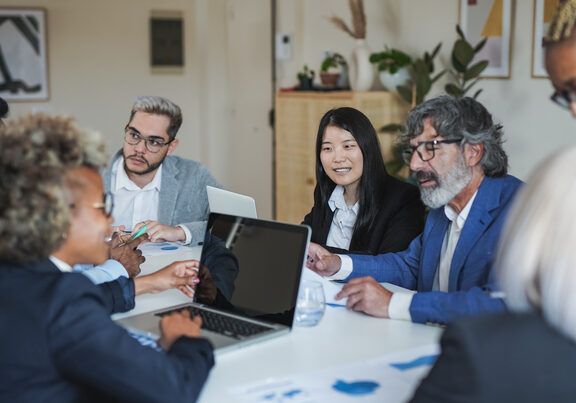 Group of people in business clothing sit around a table with laptops and paperwork